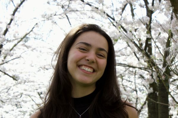 Rayane Prado Nunes, a woman with dark, long hair and wearing a sleeveless black shirt with a silver necklace. Rayane is smiling in front of numerous trees blossoming with white florals.