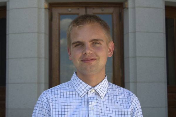 Ronan Piontek, a blonde male wearing a white and blue button up shirt, smiling in front of a stone building.